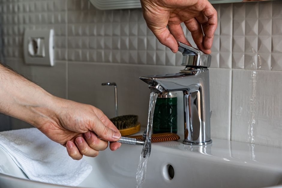 Close-up image of a man washing a shaver under a bathroom faucet, emphasizing cleanliness.
