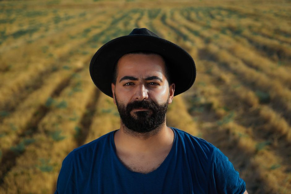 A bearded man in a blue t-shirt and hat standing in a sunlit field during summer.