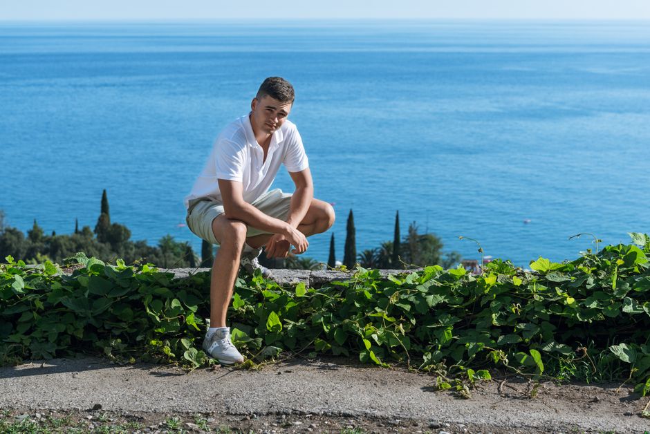 A young man crouching by the ocean, surrounded by greenery, enjoying a sunny day outdoors.