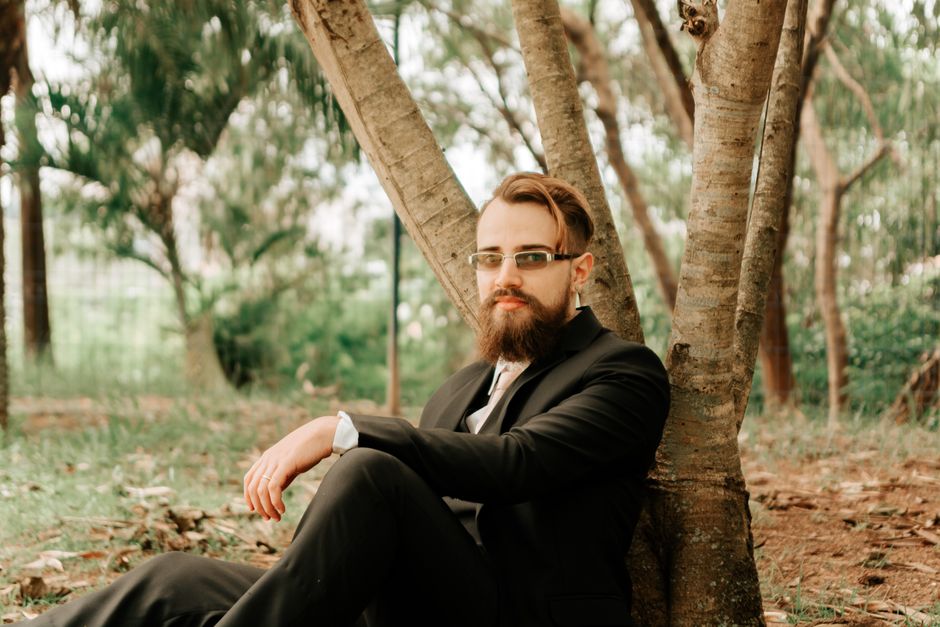 A well-dressed man with a beard sits pensively under a tree in a serene park setting.