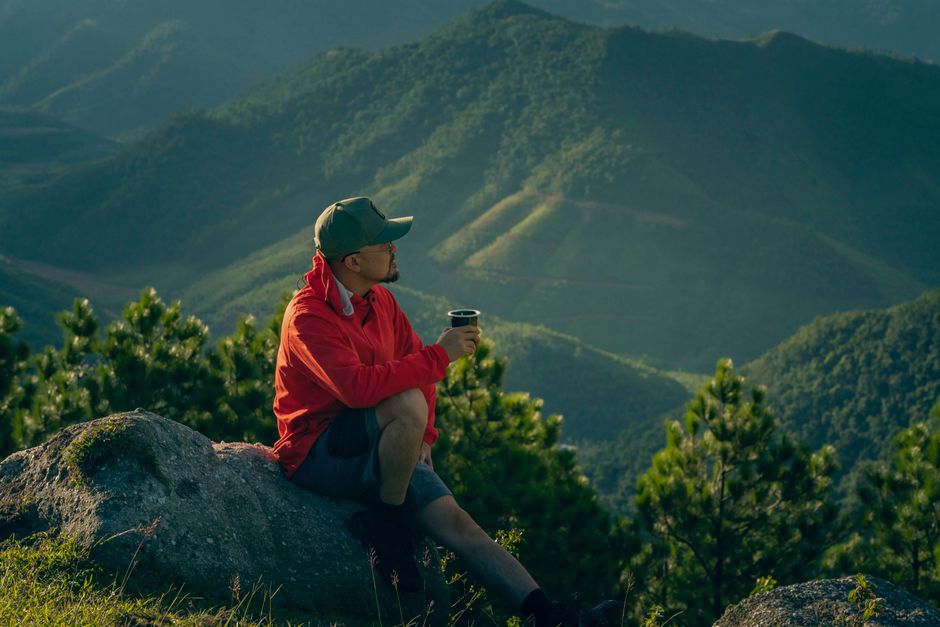 Man enjoys a serene moment in mountain scenery, sipping a drink at sunrise.