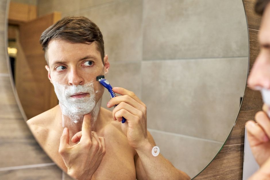 Man shaving his cheek with a razor while looking in a bathroom mirror