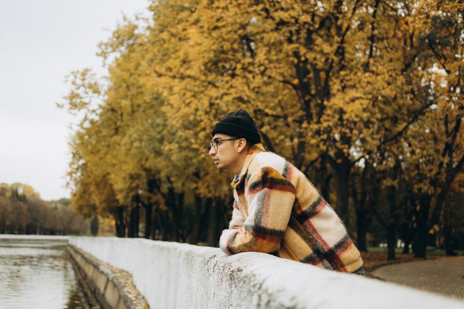 A man in a plaid jacket and beanie leans on a concrete railing with autumn trees in the background.
