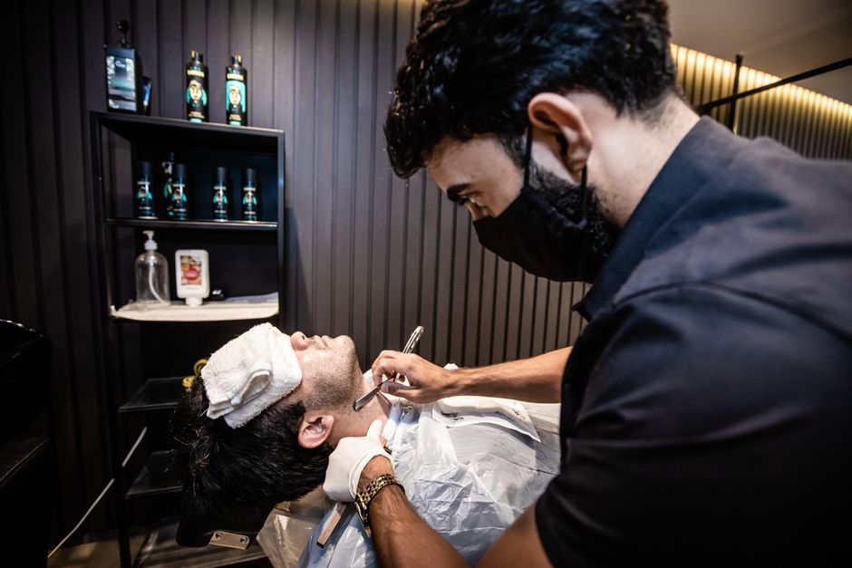 A barber carefully shaves a client's facial hair using a straight razor in a modern barbershop.