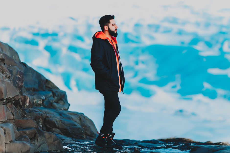 Full length of pensive ethnic man with beard wearing warm clothes looking dreamily away on blurred background of high mountains