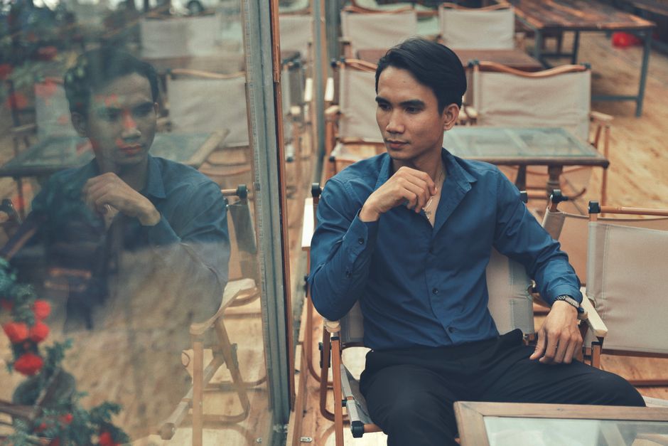 An Asian man with short black hair sitting in a café, reflecting by the window.