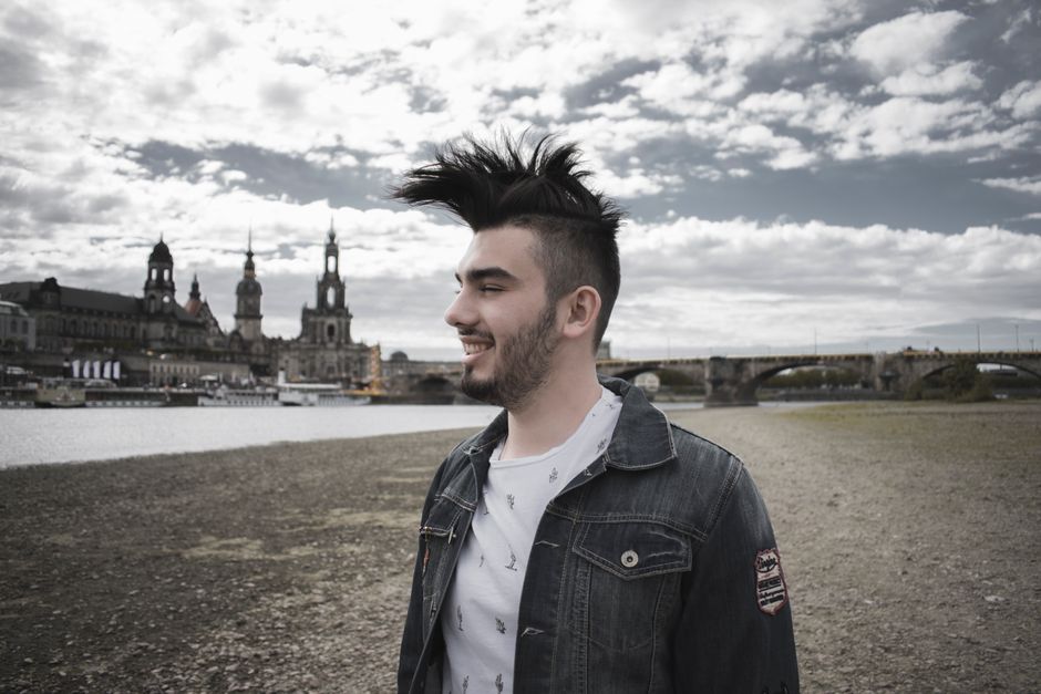 A young man with unique hairstyle smiling by the river with Dresden skyline in the background.