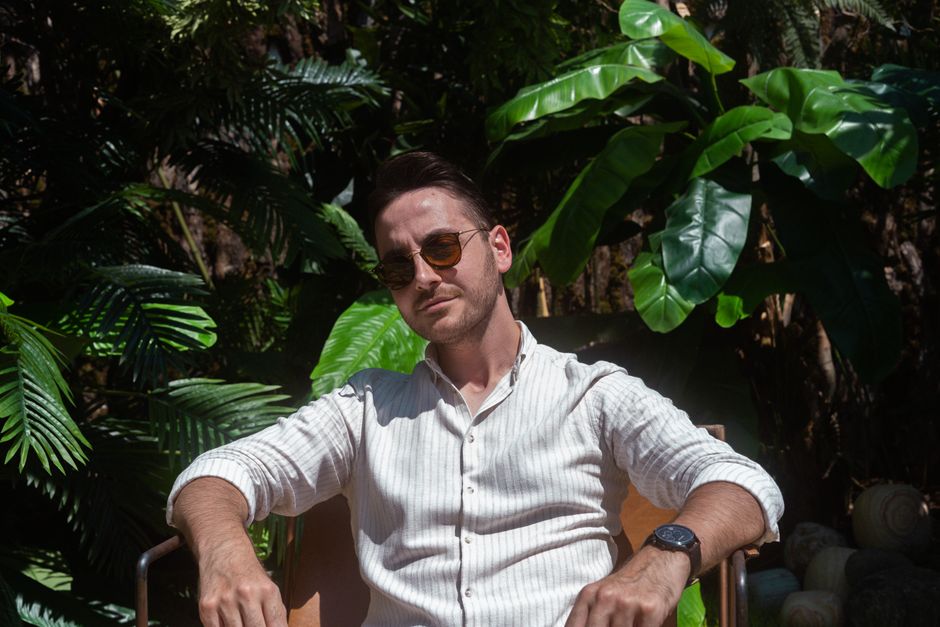 A fashionable man wearing sunglasses sits comfortably outdoors surrounded by lush greenery.