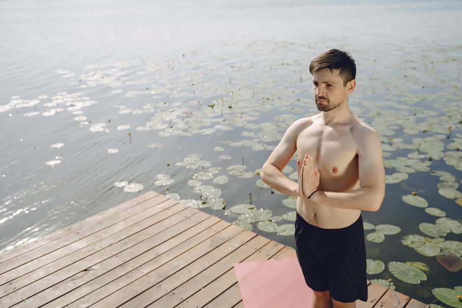A man meditating and practicing yoga on a lakeside wooden deck in summer.