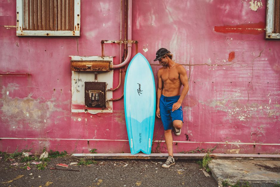 A surfer leans against a rustic pink wall with a blue surfboard, capturing the essence of Hawaii's surf culture.