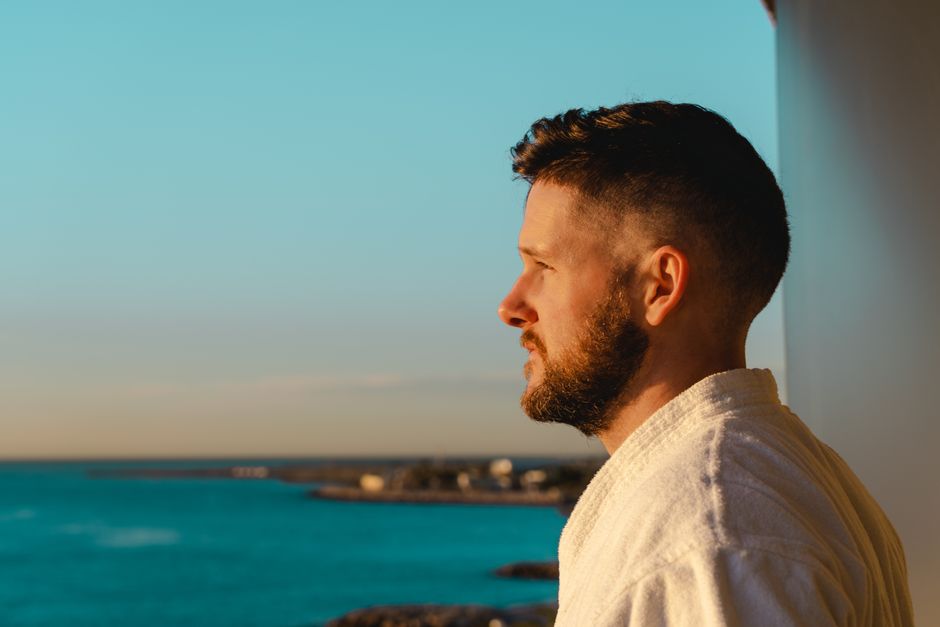 Profile of a bearded man in a white robe gazing at the ocean during sunset, capturing tranquility.