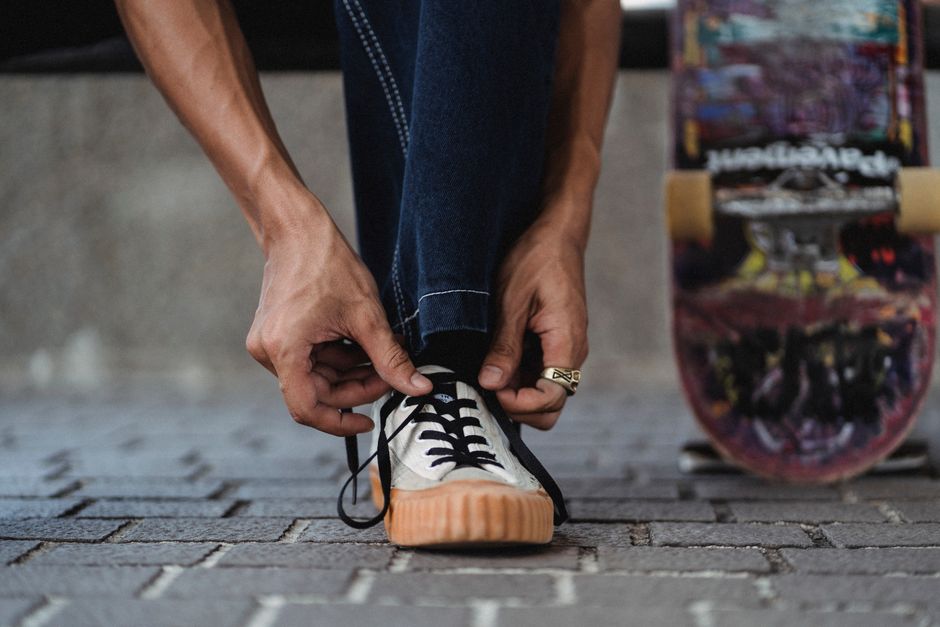 Crop unrecognizable man wearing jeans and ring on hand tying white sneakers on paved walkway with leaning colorful skateboard